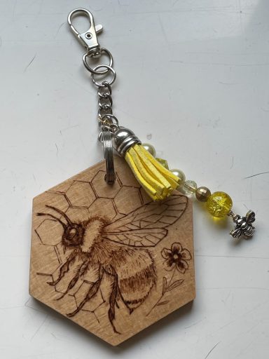 Wooden hexagonal keychain featuring a bee illustration, adorned with yellow tassel and beads.