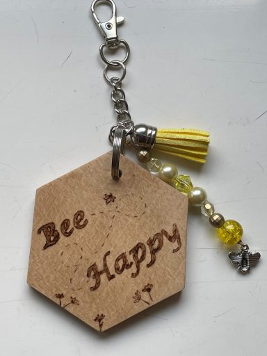 Honeycomb-shaped charm engraved with "Bee Happy," featuring a yellow tassel and beads.
